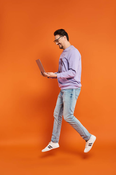 веселый человек в очках и фиолетовый свитер с помощью ноутбука на оранжевом фоне, удаленная работа - Фото, изображение