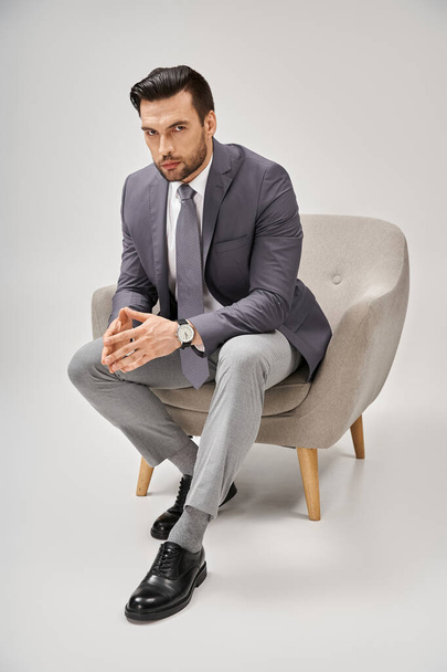 uomo d'affari sicuro e bello in abito elegante seduto su comoda poltrona su sfondo grigio - Foto, immagini