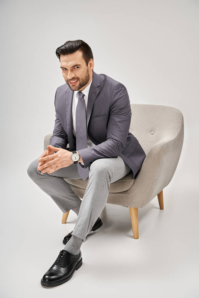 hombre de negocios alegre y guapo en traje elegante sentado en sillón cómodo sobre fondo gris - Foto, imagen