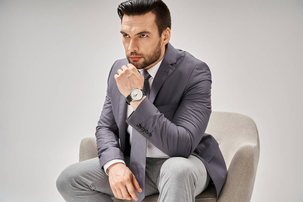 fókuszált és jóképű üzletember elegáns öltönyben ül kényelmes karosszéken szürke háttéren - Fotó, kép