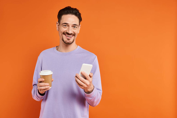 sorrindo homem em óculos segurando café para ir e usando smartphone no fundo laranja, cafeína - Foto, Imagem