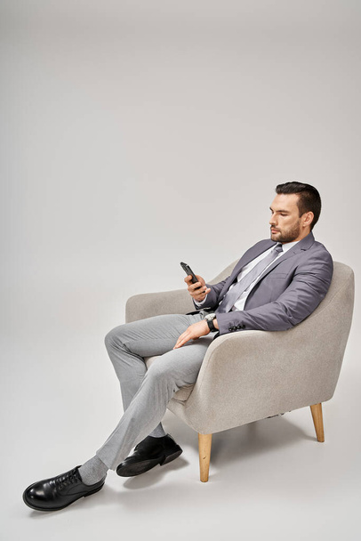 χαλαρός επιχειρηματίας που κάθεται στην πολυθρόνα και χρησιμοποιεί smartphone σε γκρι φόντο, μέσα κοινωνικής δικτύωσης - Φωτογραφία, εικόνα