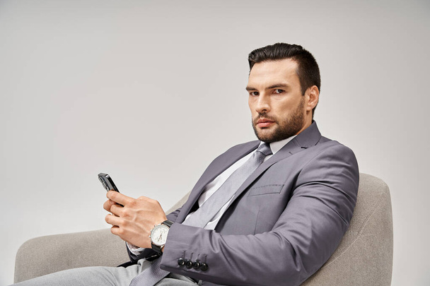 бізнесмен з щетиною сидить на кріслі і використовує смартфон на сірому фоні, підключений - Фото, зображення