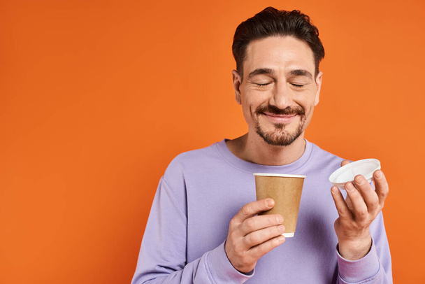 cheerful man with beard holding paper cup with coffee to go on orange background, enjoyment - Photo, Image