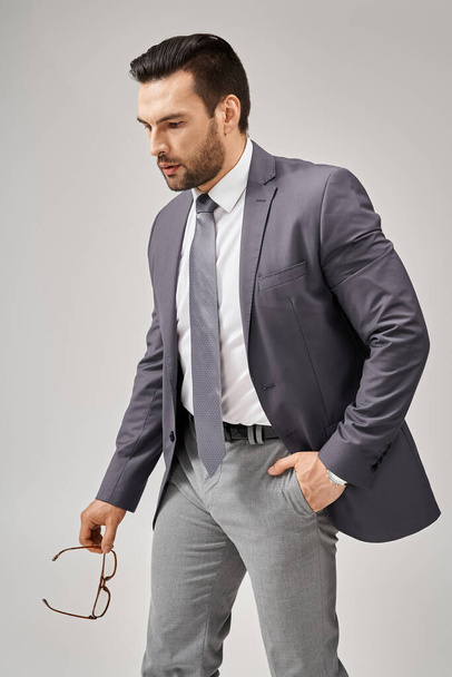 hombre de negocios equilibrado en ropa formal sosteniendo gafas y caminando con la mano en el bolsillo sobre fondo gris - Foto, Imagen