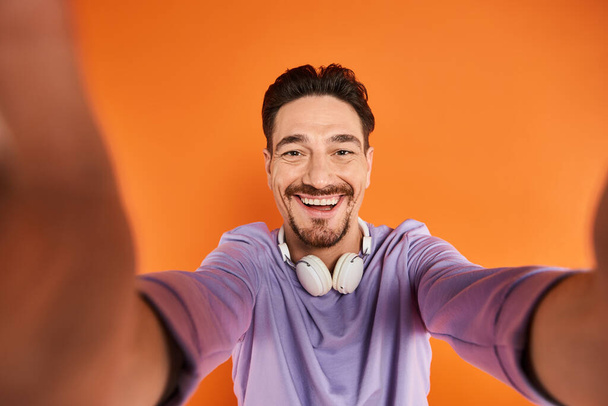 hombre barbudo feliz en auriculares inalámbricos escuchando música y mirando a la cámara en fondo naranja - Foto, imagen
