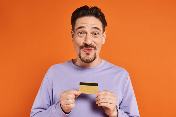vrolijk man met baard glimlachen en houden credit card op oranje achtergrond, op zoek naar camera - Foto, afbeelding
