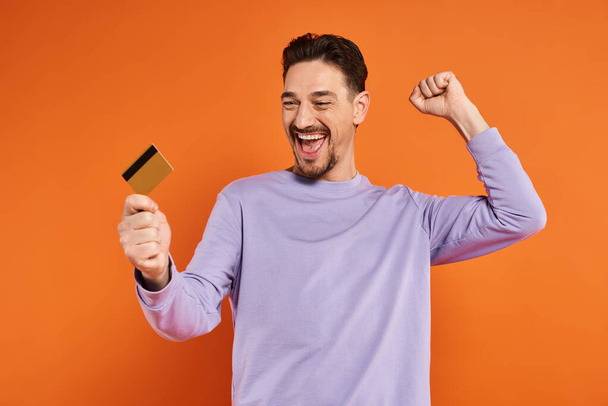 opgewonden man met baard glimlachend en met creditcard op oranje achtergrond, vrolijk gebaar - Foto, afbeelding
