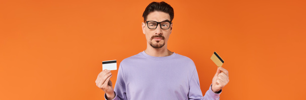 sceptische man in bril en paarse trui vergelijkt twee creditcards op oranje achtergrond, banner - Foto, afbeelding