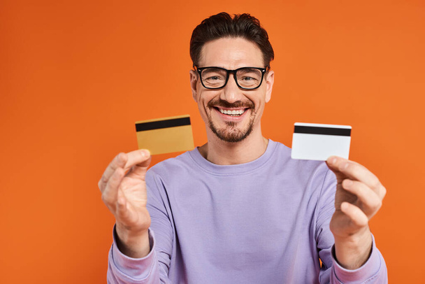 veselý vousatý muž v brýlích držící kreditní karty na oranžovém pozadí, nakupování a konzumace - Fotografie, Obrázek