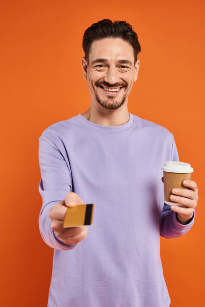 hombre feliz en suéter púrpura sosteniendo café para ir y ofreciendo tarjeta de crédito sobre fondo naranja - Foto, Imagen