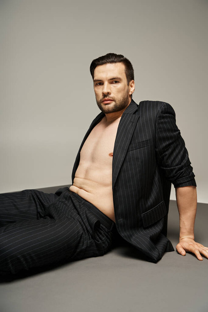 портрет стильного і красивого чоловіка з голими грудьми, що позує в одязі зі стрічкою на сірому фоні - Фото, зображення
