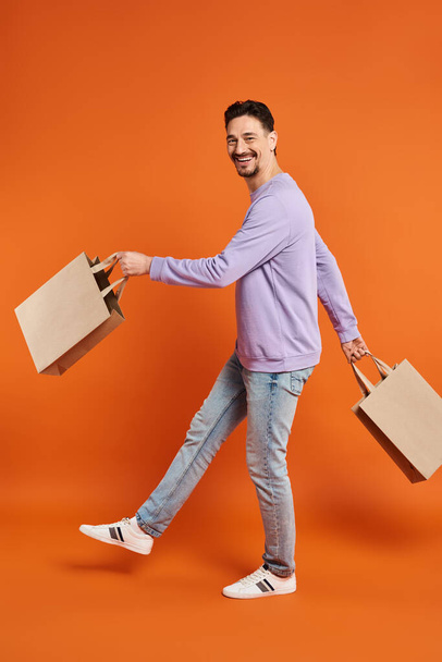 full length of happy bearded man in casual attire walking with shopping bags on orange background - Photo, Image