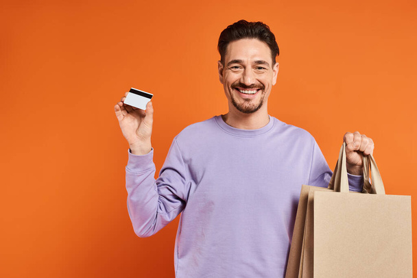 positieve bebaarde man in casual kleding houden boodschappentassen en credit card op oranje achtergrond - Foto, afbeelding