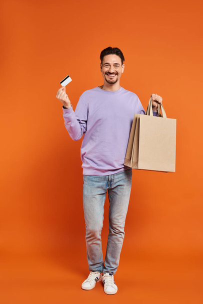 веселий бородатий чоловік у повсякденному вбранні тримає сумки і кредитну картку на помаранчевому фоні - Фото, зображення