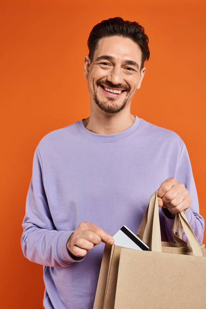 vrolijk baard man in casual kleding zetten credit card in winkeltassen op oranje achtergrond - Foto, afbeelding