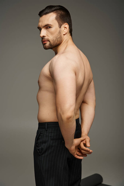 Портрет красивого мужчины без рубашки с щетиной и обнаженной грудью, позирующего в брюках на сером фоне - Фото, изображение
