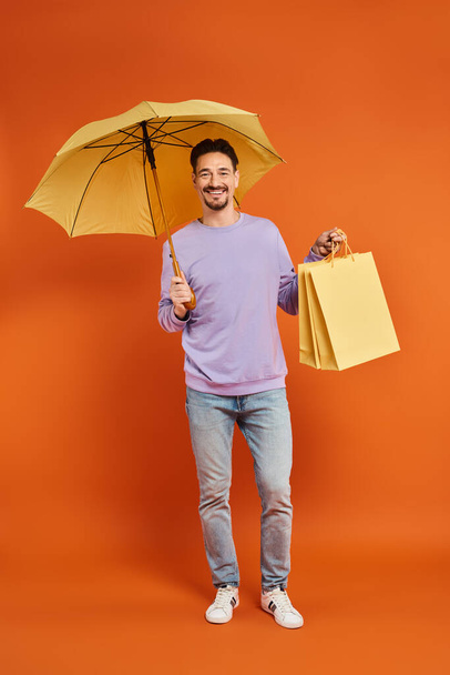 homem feliz com guarda-chuva amarelo e sacos de compras em pé sobre fundo laranja, consumismo - Foto, Imagem