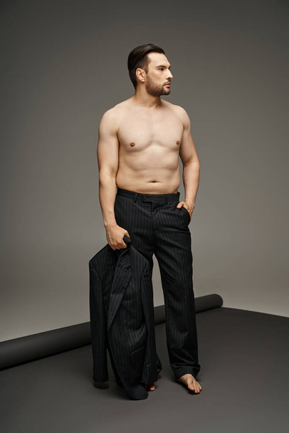 брюнетка чоловік в його 30-х позує з голими грудьми в костюмі шпильки і тримає блейзер, рука в кишені - Фото, зображення