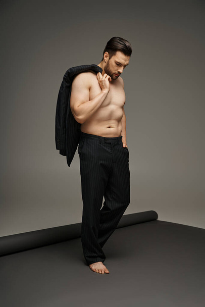 Όμορφος άντρας γύρω στα 30 ποζάροντας με γυμνό στήθος με ριγέ κοστούμι και κρατώντας σακάκι, χέρι στην τσέπη - Φωτογραφία, εικόνα