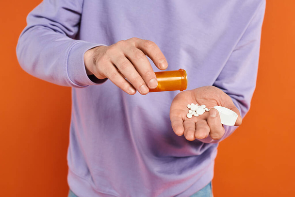 man met baard in paars sweatshirt gieten pillen in de hand op oranje achtergrond, medicatie - Foto, afbeelding