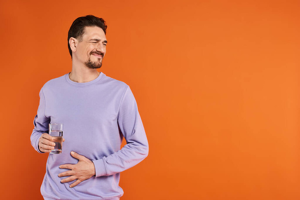bärtiger Mann in lila Sweatshirt mit Bauchschmerzen und einem Glas Wasser auf orangefarbenem Hintergrund - Foto, Bild