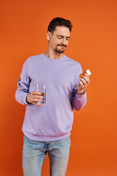 hombre barbudo en sudadera púrpura sosteniendo vaso de agua y botella con pastillas sobre fondo naranja - Foto, imagen