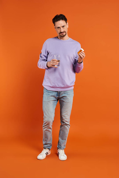 bearded man in purple sweatshirt holding glass of water and bottle with pills on orange background - Photo, Image