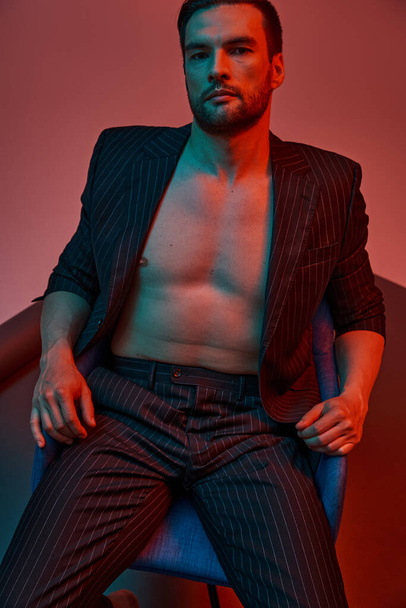 brunette man met blote borst poseren in pinstripe pak en zitten in de studio met rood en blauw licht - Foto, afbeelding