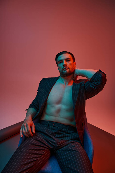 man with bare chest posing in elegant pinstripe suit and sitting in studio with red and blue light - Photo, Image