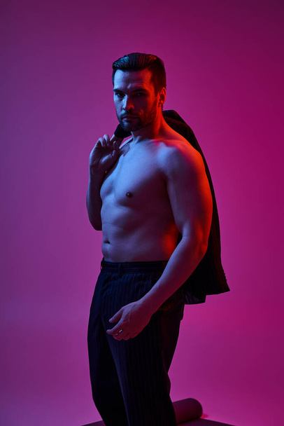 uomo muscoloso sulla trentina in posa con petto nudo in pantaloni e tenendo blazer su sfondo viola - Foto, immagini