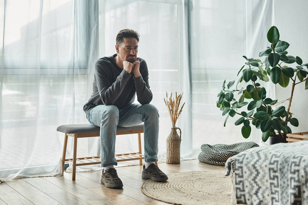 αναστατωμένος γενειοφόρος άνδρας σε casual σπίτι φορούν κάθεται στον πάγκο υπνοδωμάτιο και κοιτάζοντας μακριά, κατάθλιψη - Φωτογραφία, εικόνα