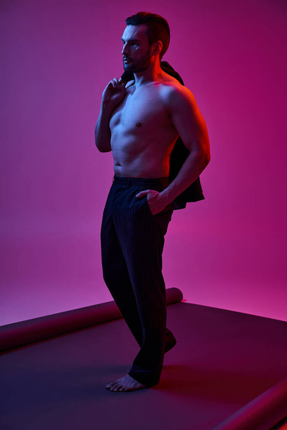 без сорочки чоловік з голими грудьми і щетиною тримає піджак, позує з рукою в кишені на фіолетовому - Фото, зображення