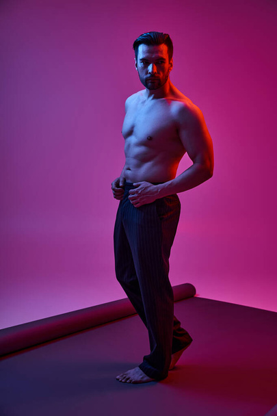 σαγηνευτικός και ημίγυμνος άντρας ποζάρει με ριγέ παντελόνι σε μωβ φόντο με κόκκινα και μπλε φώτα - Φωτογραφία, εικόνα