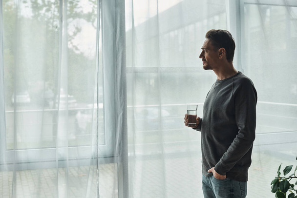 вдумчивый бородатый мужчина, стоящий с рукой в кармане, глядя в окно и держа стакан воды - Фото, изображение