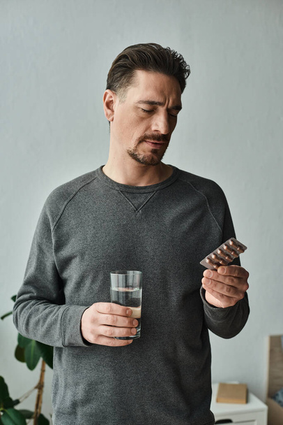 uomo barbuto in casual maglione grigio aggrottare le sopracciglia e tenere in mano un bicchiere d'acqua mentre guardando il farmaco - Foto, immagini