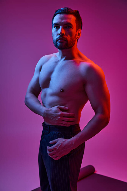 schöner Mann mit nackter Brust posiert in Nadelstreifenhosen vor lila Hintergrund mit rotem Licht - Foto, Bild