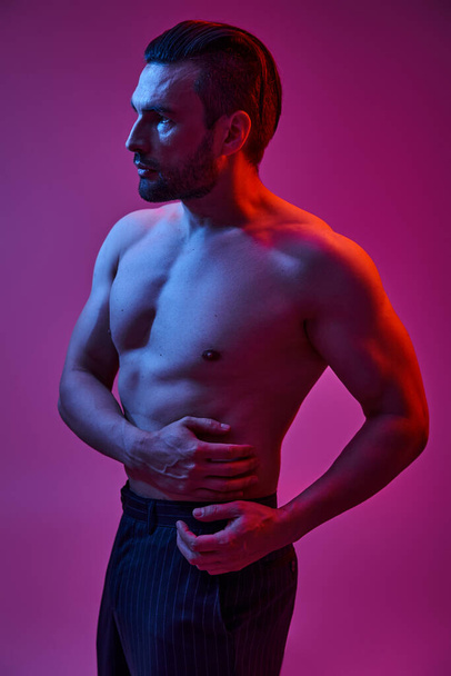 svůdný muž s nahou hrudí pózující v proužkovaných kalhotách na purpurovém pozadí s červenými světly - Fotografie, Obrázek