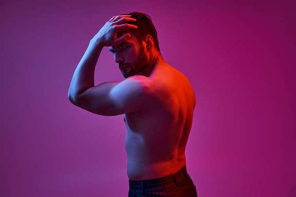 спокусливий чоловік з сексуальними голими грудьми позує рукою біля волосся на фіолетовому тлі з червоними вогнями - Фото, зображення