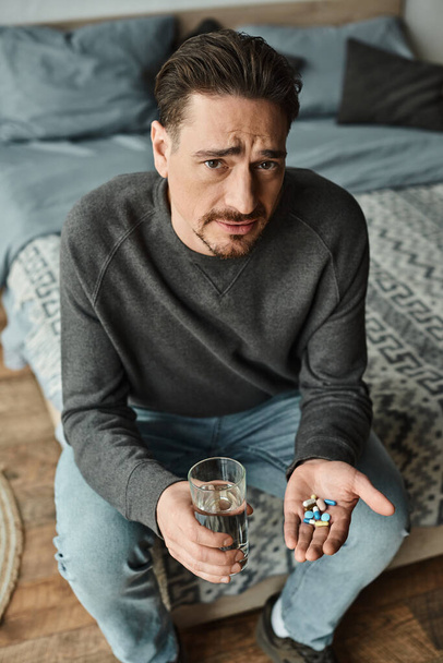 szakállas férfi ráncolja homlokát, miközben egy pohár vizet és gyógyszert tart a kezében, a kamerába néz. - Fotó, kép
