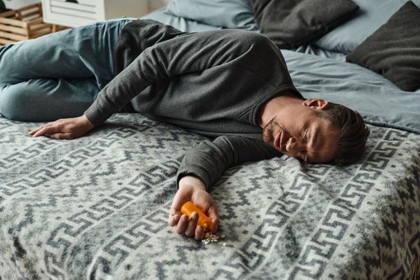 бородатий чоловік страждає від болю і дивиться на пляшку з таблетками, лежачи на ліжку з ковдрою - Фото, зображення
