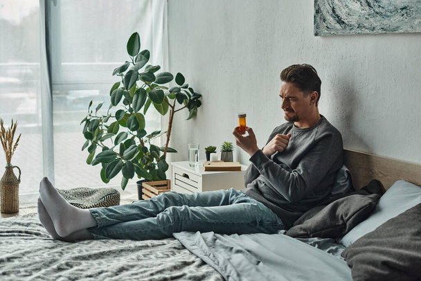 hombre pensativo examinando botella con medicamentos mientras está acostado en la cama en el dormitorio, plan de tratamiento - Foto, Imagen