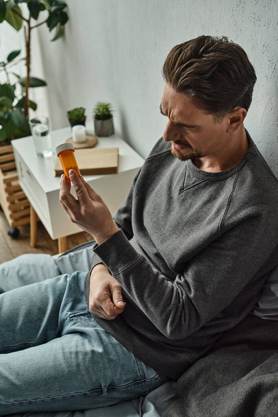 töprengő férfi vizsgálja üveg gyógyszert, miközben ül az ágyon a hálószobában, kezelési terv - Fotó, kép