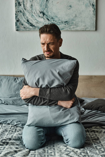 man met baard die zich ongemakkelijk voelt en een kussen vasthoudt terwijl hij thuis op bed zit, patiënt in moeilijkheden - Foto, afbeelding