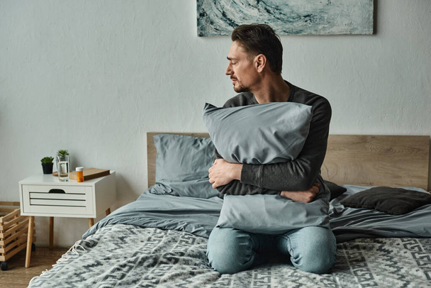 man met baard die zich ongemakkelijk voelt en een kussen vasthoudt terwijl hij thuis op bed zit, modern interieur - Foto, afbeelding