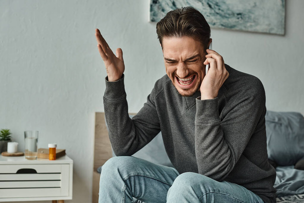 Wütender bärtiger Mann im grauen Pullover, der im modernen Schlafzimmer mit dem Smartphone spricht und gestikuliert - Foto, Bild