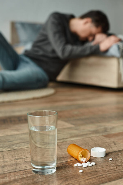 стакан воды и бутылка с таблетками на полу возле размытого человека, страдающего от боли на заднем плане - Фото, изображение