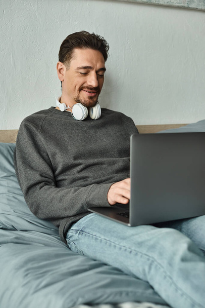 χαρούμενος άνθρωπος σε ασύρματα ακουστικά χρησιμοποιώντας φορητό υπολογιστή, ενώ κάθεται στο κρεβάτι, freelance και απομακρυσμένη εργασία - Φωτογραφία, εικόνα
