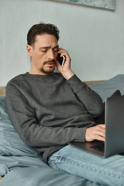 Sakallı adam bilgisayarını kullanıyor ve evden uzakta çalışırken cep telefonuyla konuşuyor. - Fotoğraf, Görsel