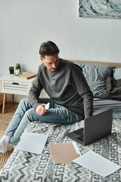 сфокусированный бородатый мужчина держит смартфон и использует ноутбук рядом с документами на кровати, удаленная работа - Фото, изображение